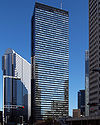 Ground view of a black building’s two rectangular, reflective glass facades set in front of a dark blue sky and surrounded by other tall buildings; the smaller facade is bisected by black, inset, crisscrossed beams