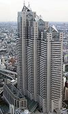 Aerial view of a building’s two beige facades with horizontal rows of windows set in front of a cityscape; the building is composed of three adjoined towers of differing heights, each capped off with triangular glass structures