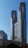 Ground-level view of a grey, window-dotted front building facade set in front of a dark blue sky; as the building rises, two towers break off on both sides